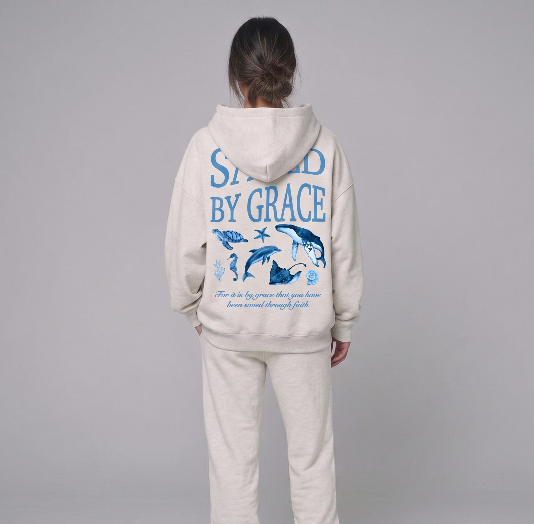 Person wearing a beige hoodie and pants set with 'Saved by Grace' text on the back, on a gray background.